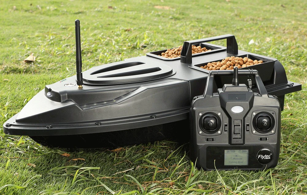 Barcă de momeală RC neagră cu două recipiente pentru pelete și o telecomandă Flytec așezată pe iarbă