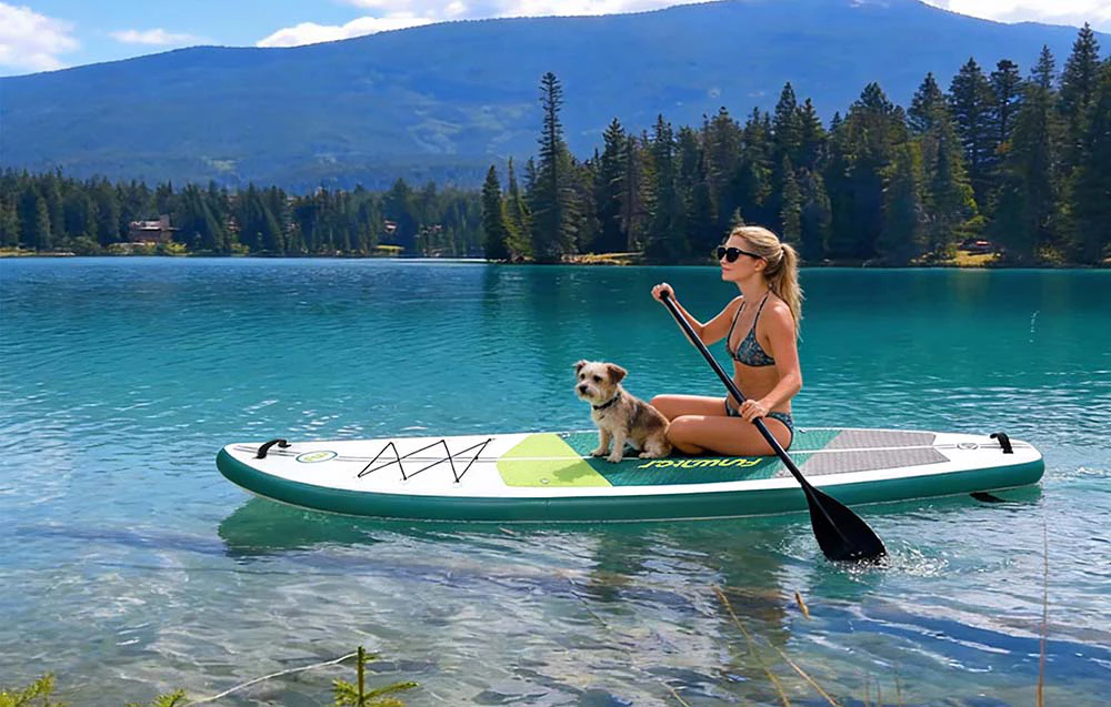 O femeie în costum de baie și ochelari de soare stă pe o placă de surf ținând o vâslă în mână. Un câine stă lângă ea, cu munții în fundal