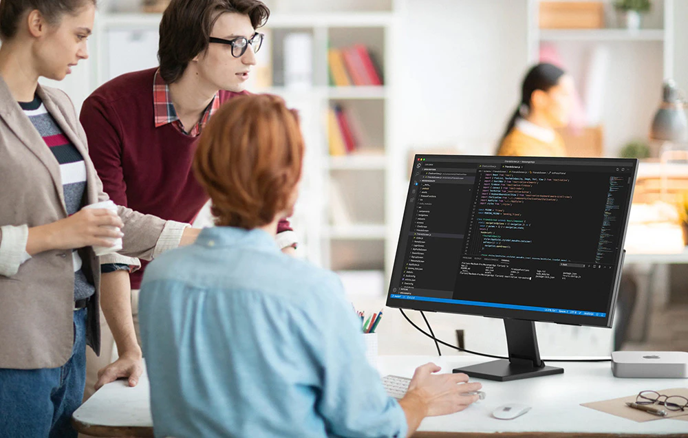 Trei persoane într-un birou lucrând la un monitor care afișează un editor de cod