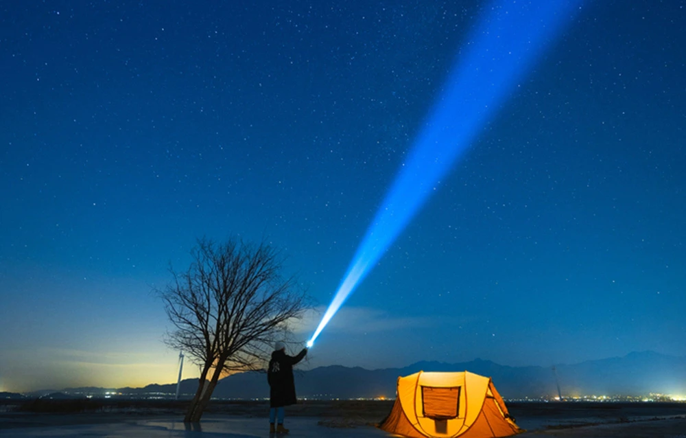 Silueta unei persoane cu o lanternă care emite o rază de lumină albastră spre cer lângă un cort portocaliu