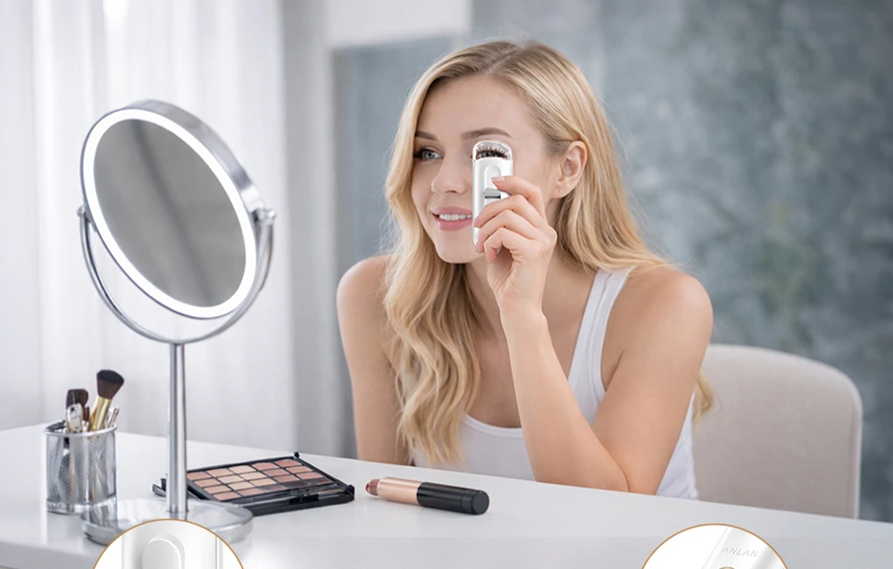 O femeie folosește un ondulator de gene alb în fața unei oglinzi cosmetice iluminate