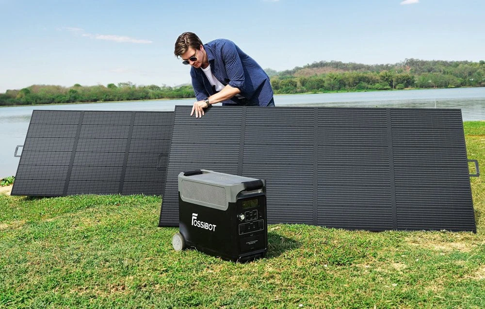 Un bărbat desfășoară un panou solar Fossibot de mari dimensiuni lângă un lac, cu o stație de alimentare portabilă amplasată în apropiere