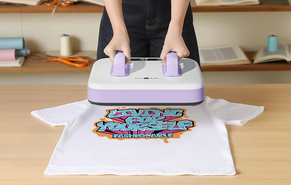 O persoană ține mânerele violet ale presei termice apăsate pe un tricou alb cu litere colorate.