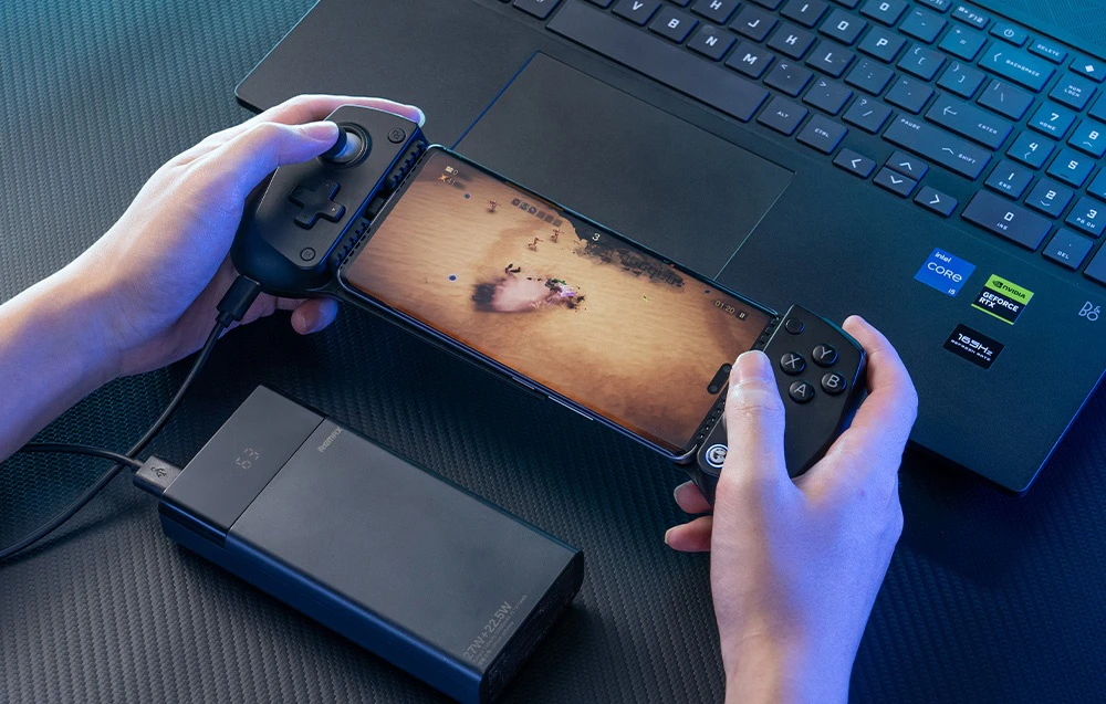 O persoană care joacă pe un smartphone montat într-un controller GameSir negru cu un powerbank conectat lângă un laptop.