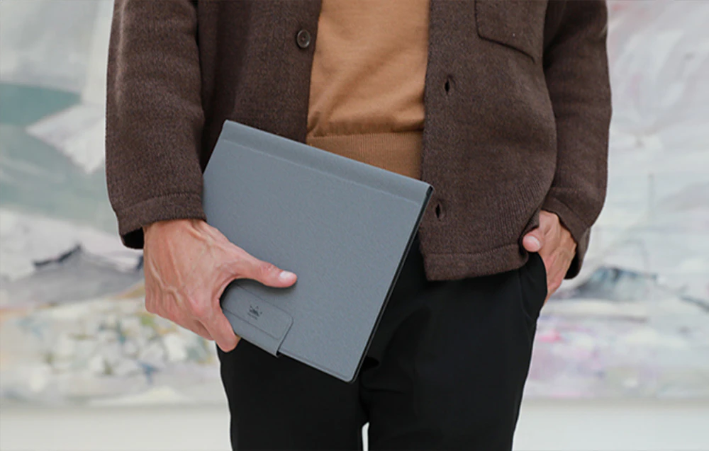 O persoană într-un pulover maro ține sub braț o tabletă Huion închisă într-o carcasă gri.