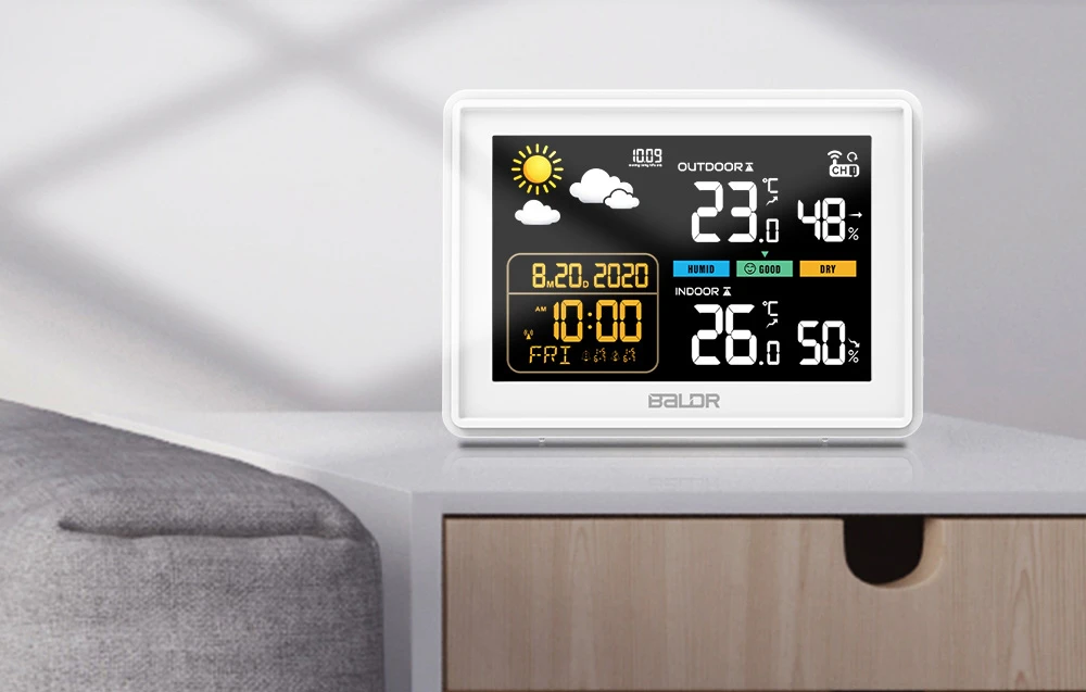 O stație meteorologică electronică cu ecran LCD color care afișează temperatura, umiditatea și ora, așezată pe o comodă albă.