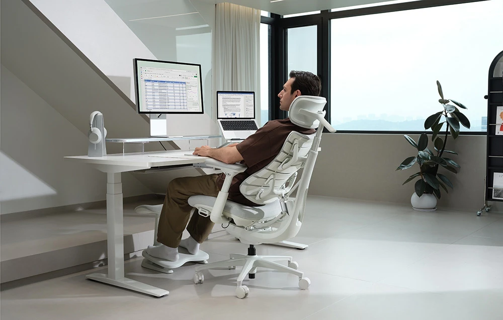 Un bărbat îmbrăcat într-un tricou maro stă așezat într-un scaun ergonomic alb, la un birou alb cu un monitor