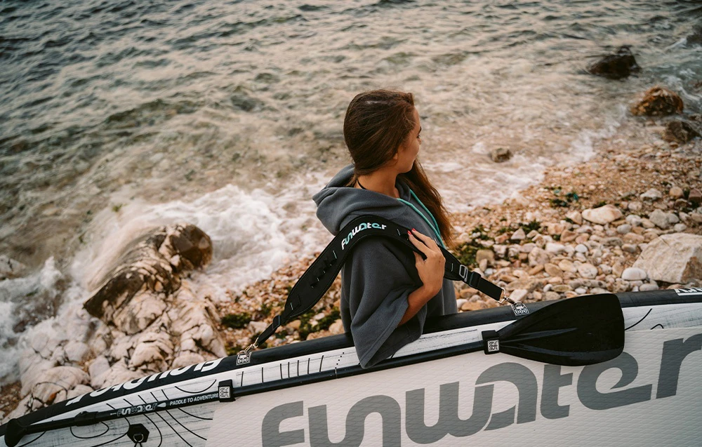 Imagine cu o femeie care transportă o placă de surf pe umăr, cu o plajă stâncoasă și apă vizibile în fundal