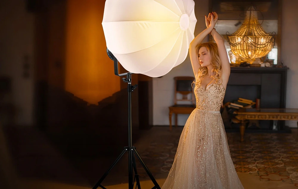 O femeie în rochie pozează lângă o lampă cu un abajur alb mare care emite lumină caldă.