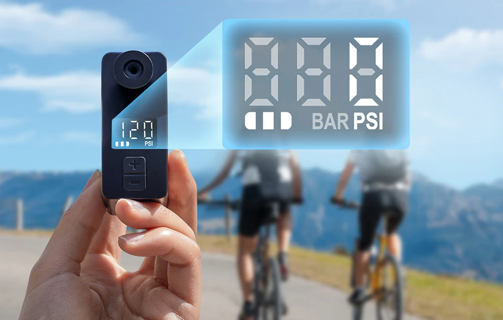 O mini-pompă de bicicletă ținută în mână pe fundalul unor cicliști, cu un manometru digital vizibil.
