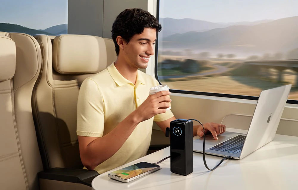 Un bărbat zâmbitor într-un tricou galben stă în tren și ține în mână o cană. Pe masa din fața sa se află un laptop și un smartphone conectat la powerbank.