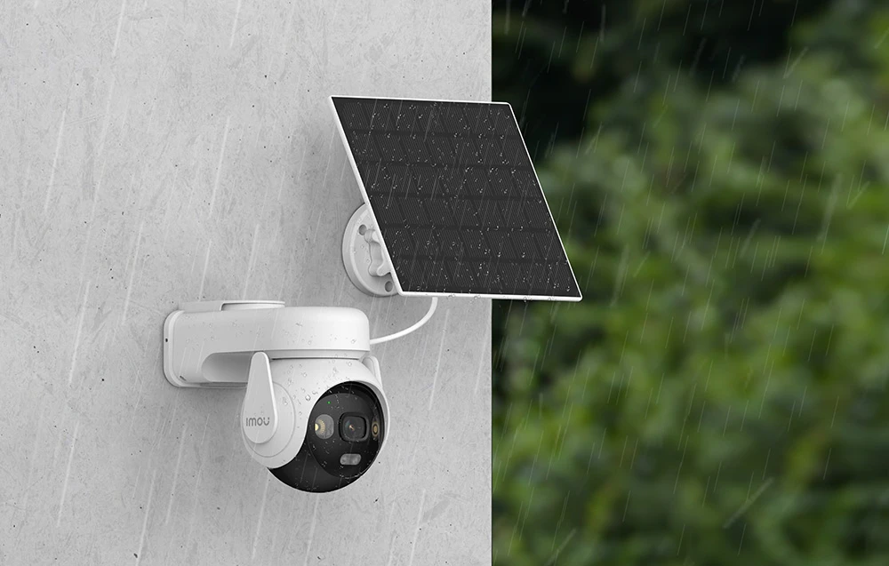 Cameră albă cu funcție pan-tilt și panou solar montată pe un perete gri în ploaie, cu copaci verzi în fundal