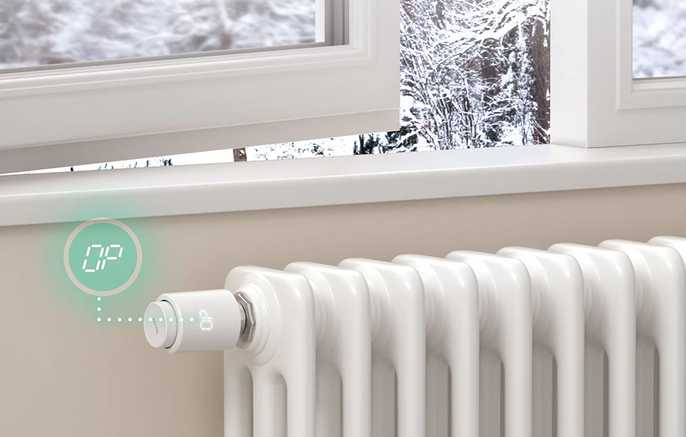 Un radiator cu un cap termostatic inteligent, o fereastră deschisă deasupra acestuia și un peisaj de iarnă în fundal