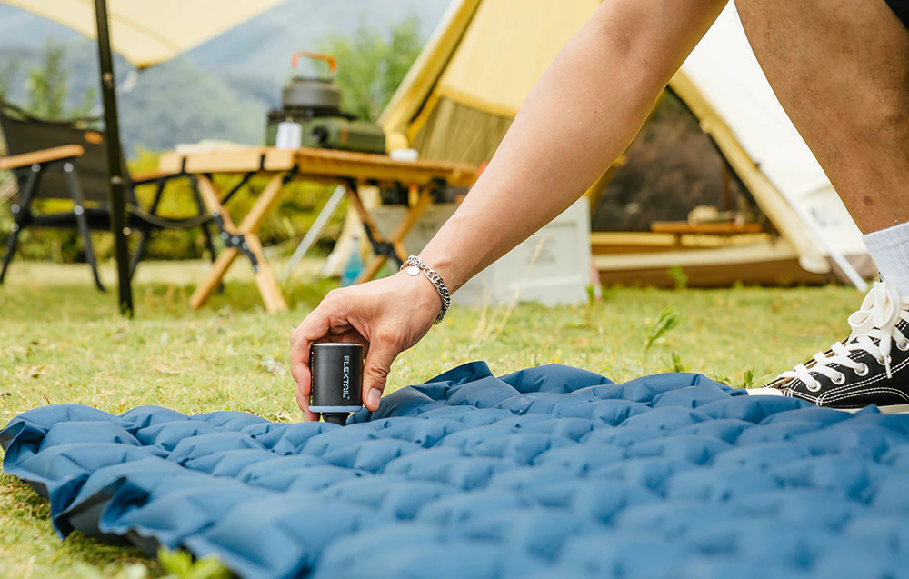 Un bărbat folosește o pompă Flextail mică, neagră, pentru a umfla un covoraș de trekking albastru într-un camping