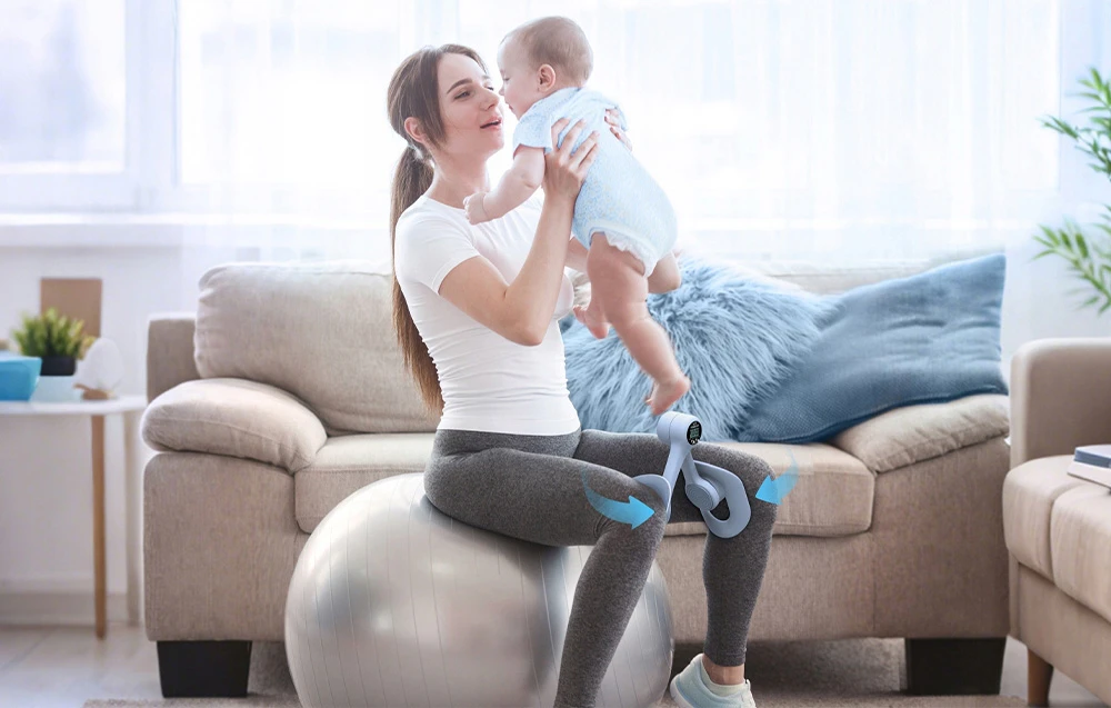 O femeie stă așezată pe o minge de exerciții, ținând un bebeluș în brațe și antrenându-și coapsele cu un dispozitiv de exerciții de culoare albastru deschis