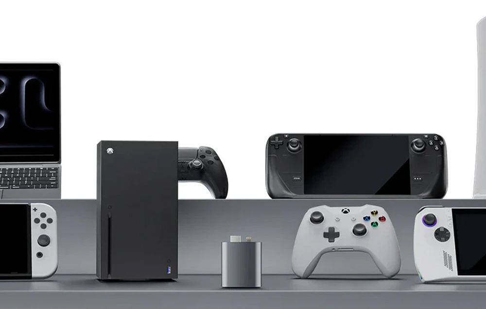 Adaptorul plasat pe un fundal cu diverse dispozitive de jocuri și multimedia, inclusiv console, controlere, laptopuri și dispozitive portabile.
