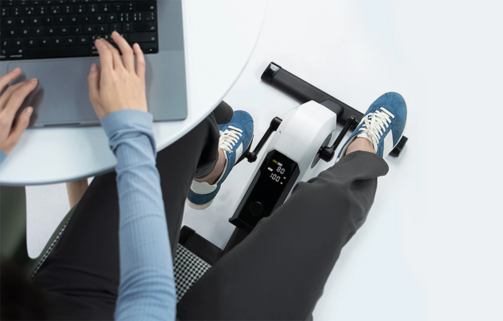 Persoană care lucrează la un laptop la o masă și folosește o bicicletă mică de reabilitare
