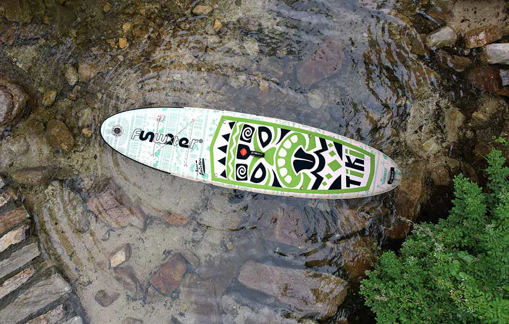 O placă de surf Funwater colorată este așezată pe apă, înconjurată de roci și plante