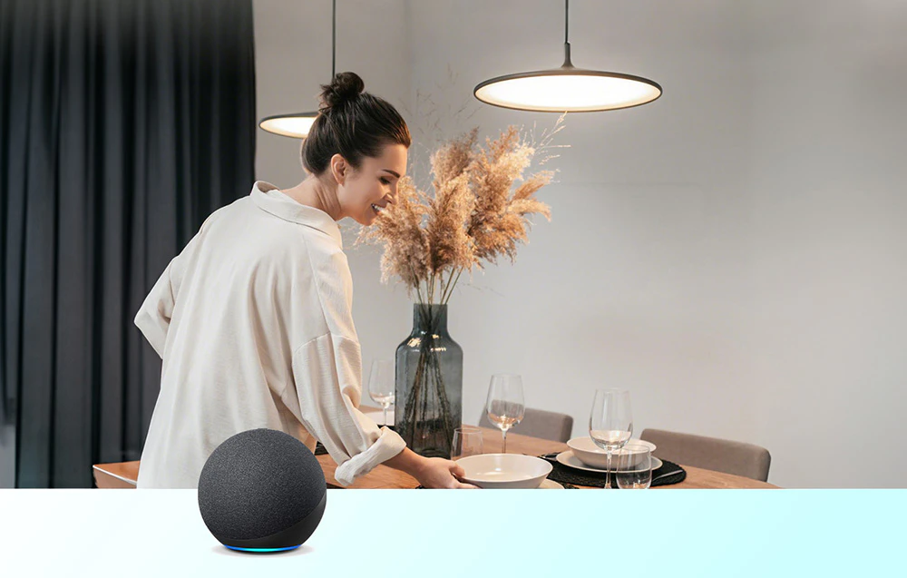 O femeie aranjează tacâmurile la o masă cu o tavă, lângă o boxă inteligentă Amazon Echo de culoare neagră aflată pe blatul mesei