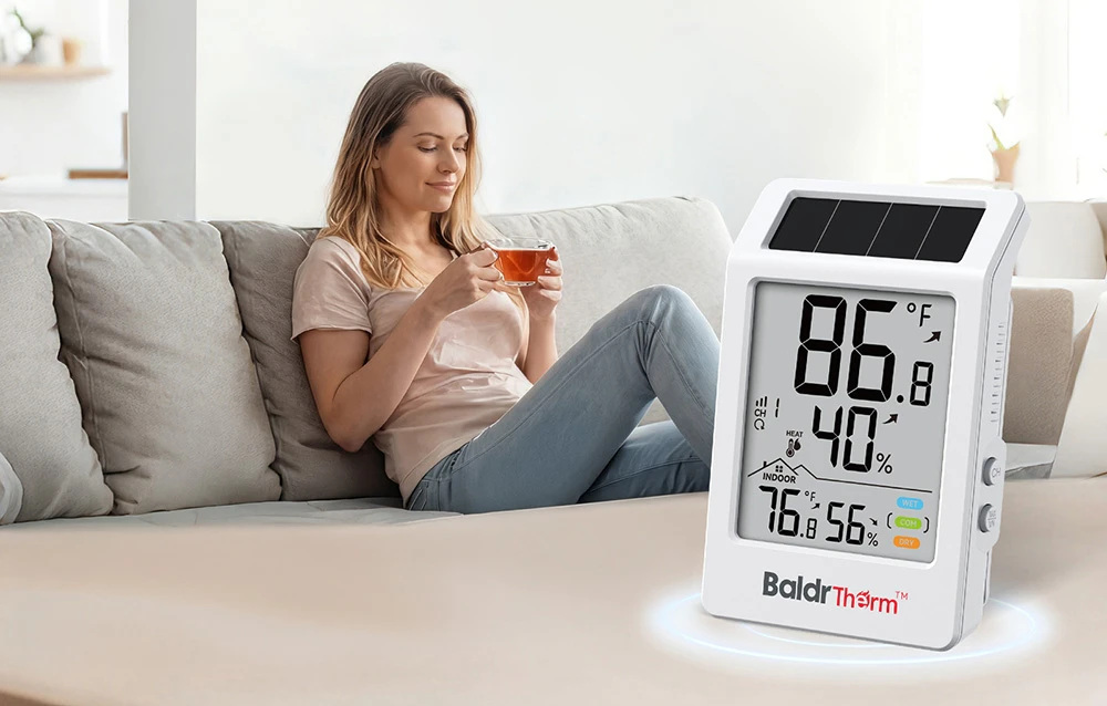 O femeie stă pe o canapea, ținând o ceașcă de ceai; un termohigrometru se află pe măsuța de cafea din prim-plan