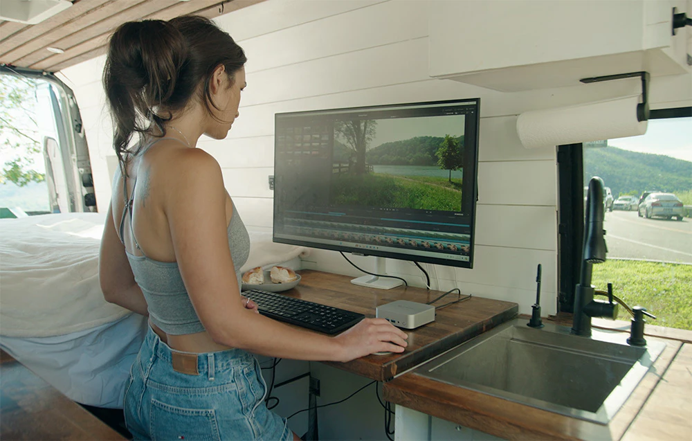 O femeie care lucrează la un mini PC în interiorul unei rulote, lângă fereastră. Pe monitor este vizibil un program de editare video.
