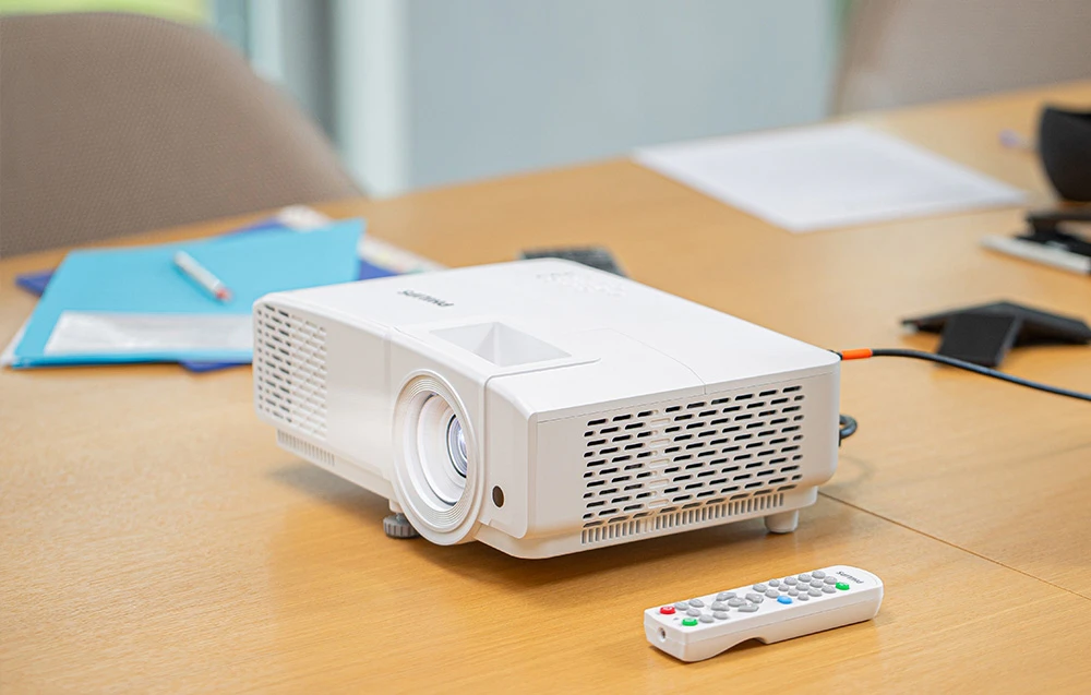 Un proiector alb PHILIPS se află pe o masă de lemn lângă o telecomandă și niște dosare albastre