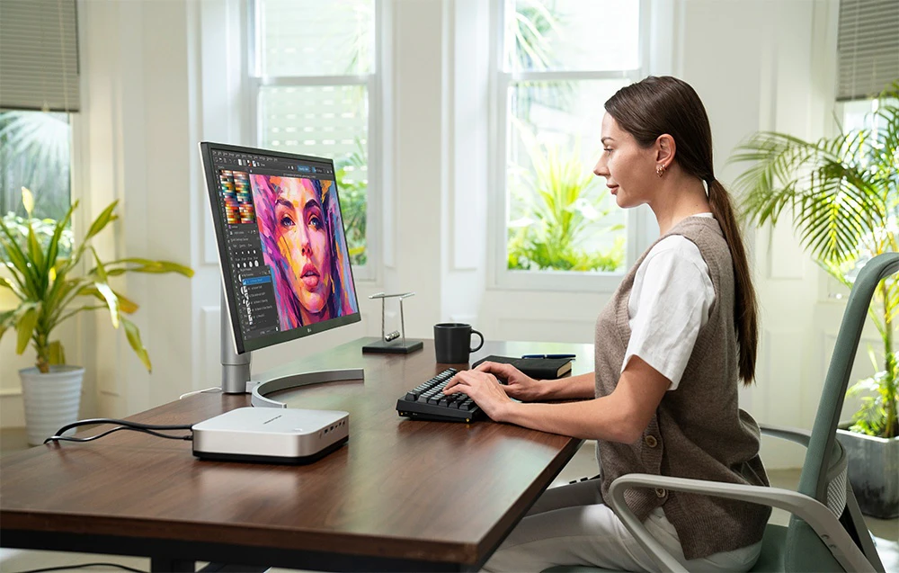 O femeie la un birou, editând grafice pe un monitor mare conectat la un Mini PC compact.