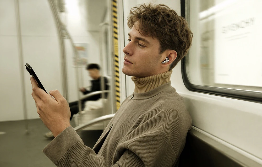 Un bărbat îmbrăcat într-un pulover cu guler înalt bej, așezat în metrou, uitându-se la ecranul telefonului, cu o cască vizibilă în ureche.