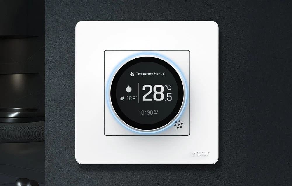 Termostat alb cu afișaj rotund indicând 28,5 °C, montat pe un perete negru