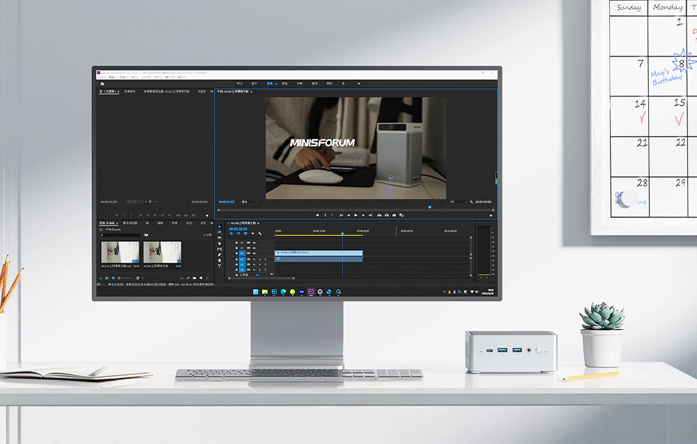 Un monitor pe un birou afișează un program de editare video, alături de un mini PC, calendar, notebook și ghiveci de flori pe un birou luminos și ordonat