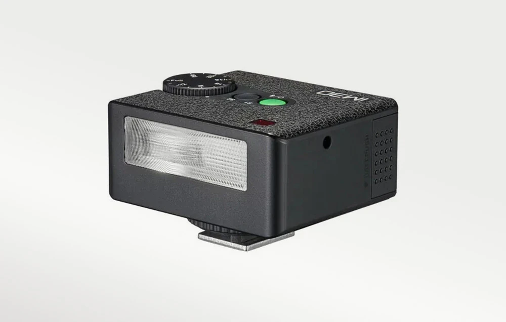 Un bliț negru cu un reflector dreptunghiular vizibil din față, o parte superioară texturată, un cadran și un picior de montare din metal.