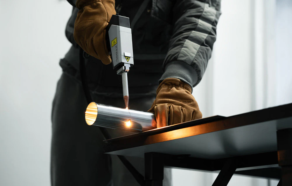 O persoană în mănuși maro sudează o țeavă metalică cu un aparat de sudură cu laser