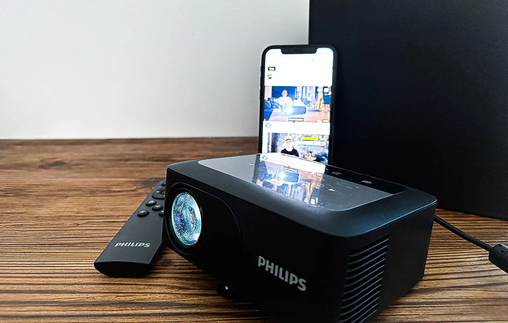 Un proiector PHILIPS negru pe o masă de lemn, cu o telecomandă PHILIPS și un smartphone care afișează un film lângă el