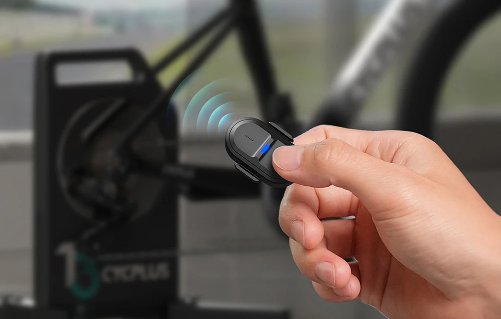 O mână care ține telecomanda wireless neagră Cycplus pentru un antrenor de bicicletă, având ca fundal o bicicletă și antrenorul de bicicletă Cycplus T2