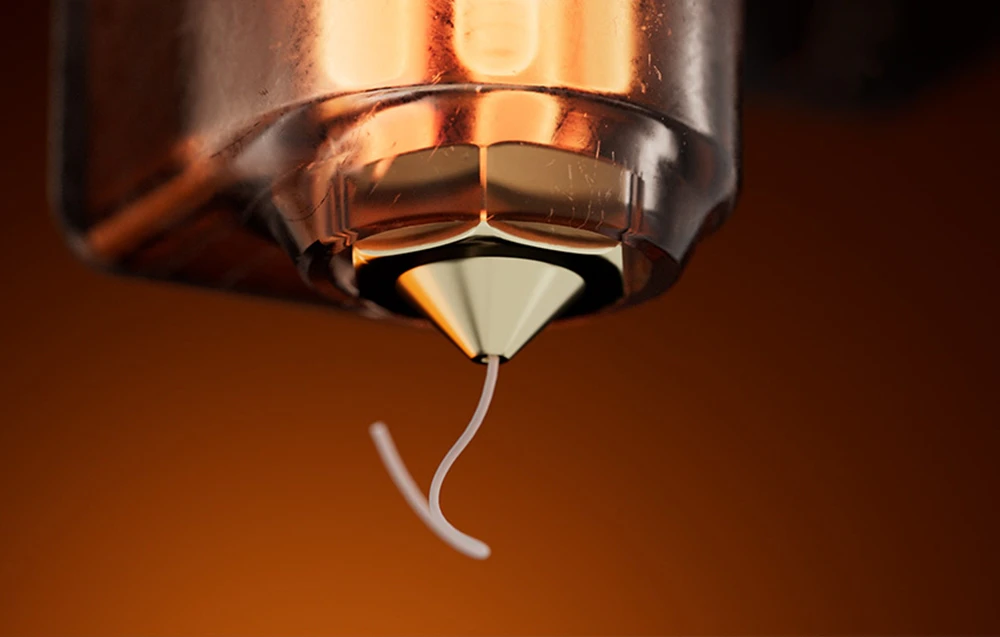 Fotografie macro a unei duze conice din alamă, din care curge un fir subțire de plastic topit.