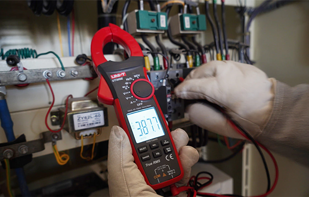 Un clește ampermetric digital UNI-T ținut de un electrician care poartă mănuși în timpul testării într-un dulap de comandă.
