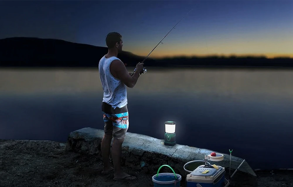 Un pescar stă pe mal, lângă o lampă de camping aprinsă și niște găleți, cu un lac în fundal.
