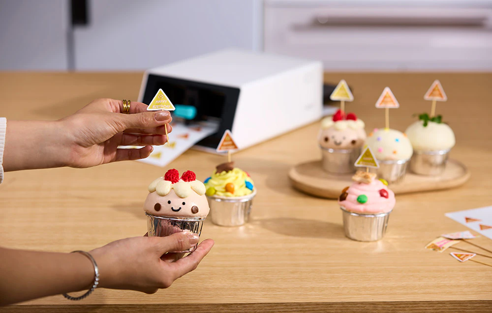 O mână așezând un steag mic cu text pe un cupcake umplut cu cremă; în fundal, imprimanta Liene pe o masă din lemn.