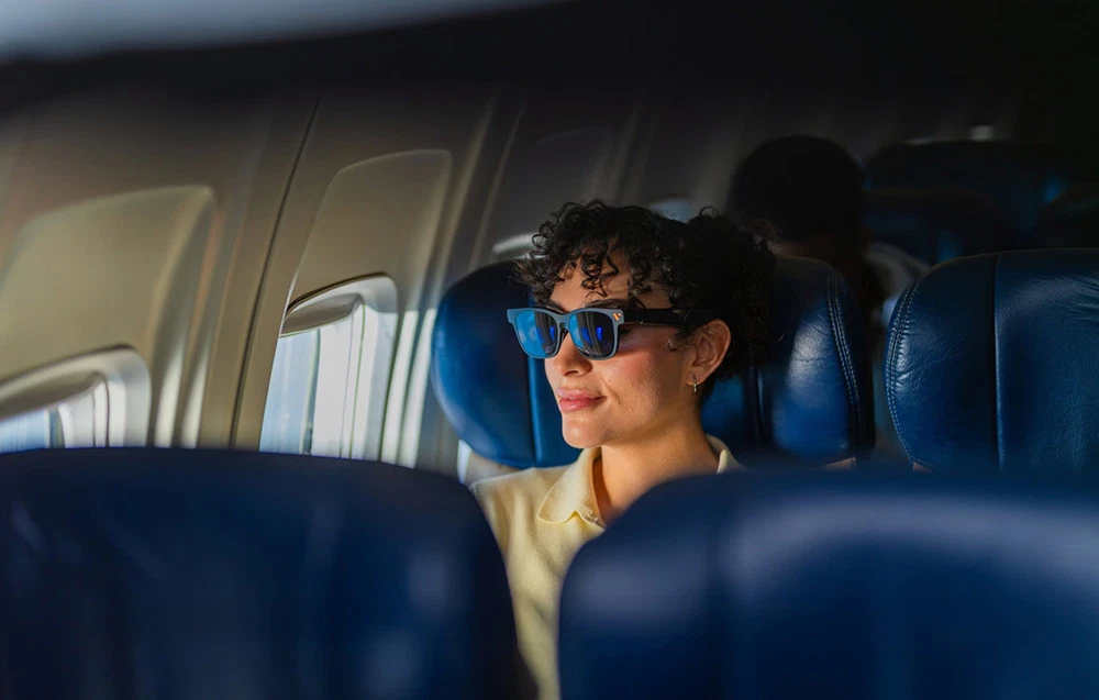 O femeie care poartă ochelari negri stă așezată într-un scaun de avion albastru