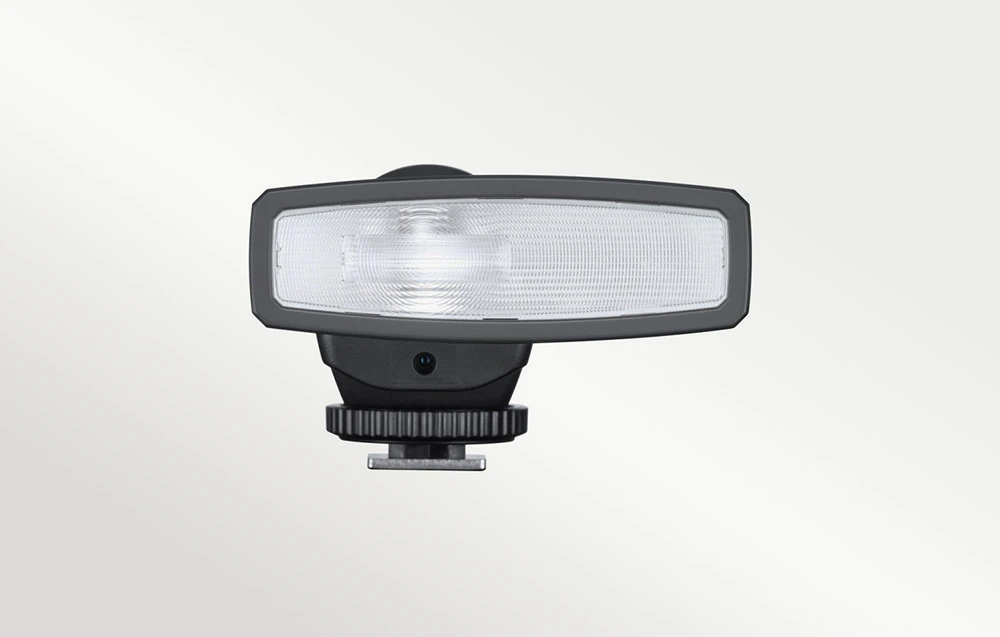 Un bliț negru cu un reflector alungit și luminos în partea din față și un hot shoe metalic pe un fundal deschis