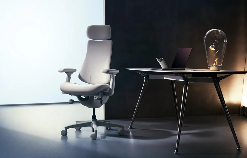 Un scaun de birou ergonomic cu tetieră, așezat lângă un birou modern cu un laptop și o lampă, într-o cameră întunecată