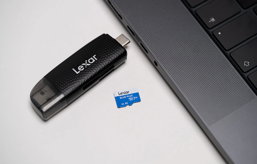 Cardul de memorie Lexar Blue Plus microSDXC așezat pe o masă albă lângă un cititor de carduri USB-C negru și marginea unui laptop gri.