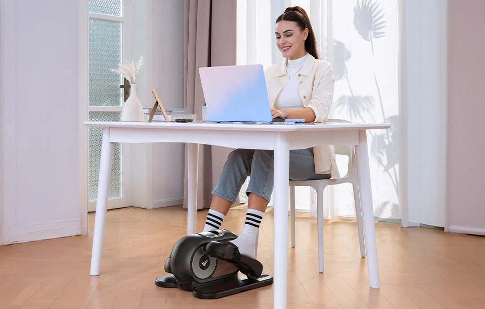 O femeie lucrează la laptopul ei la birou în timp ce utilizează mini-elipticul Merach sub picioare.