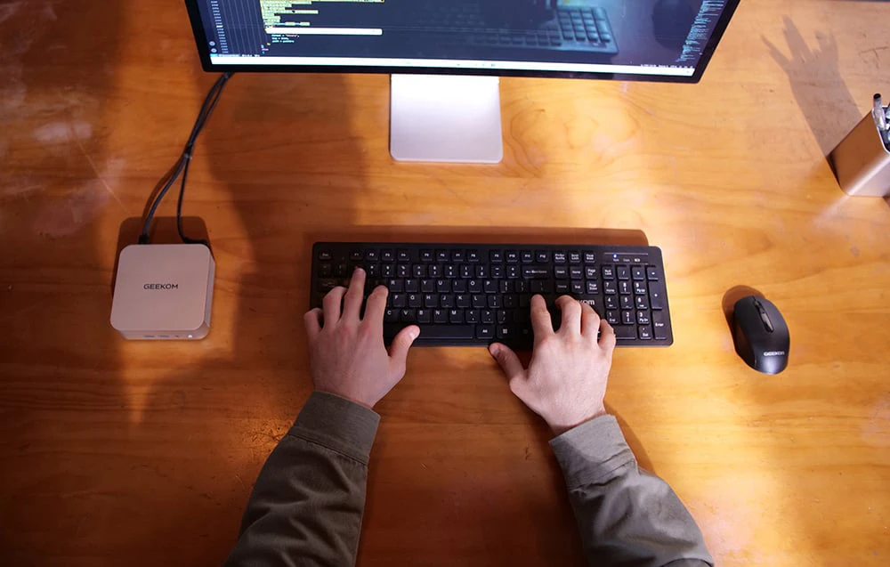 O persoană așezată la un birou și tastând la o tastatură, cu un monitor, un mouse și un mini PC Geekom în apropiere