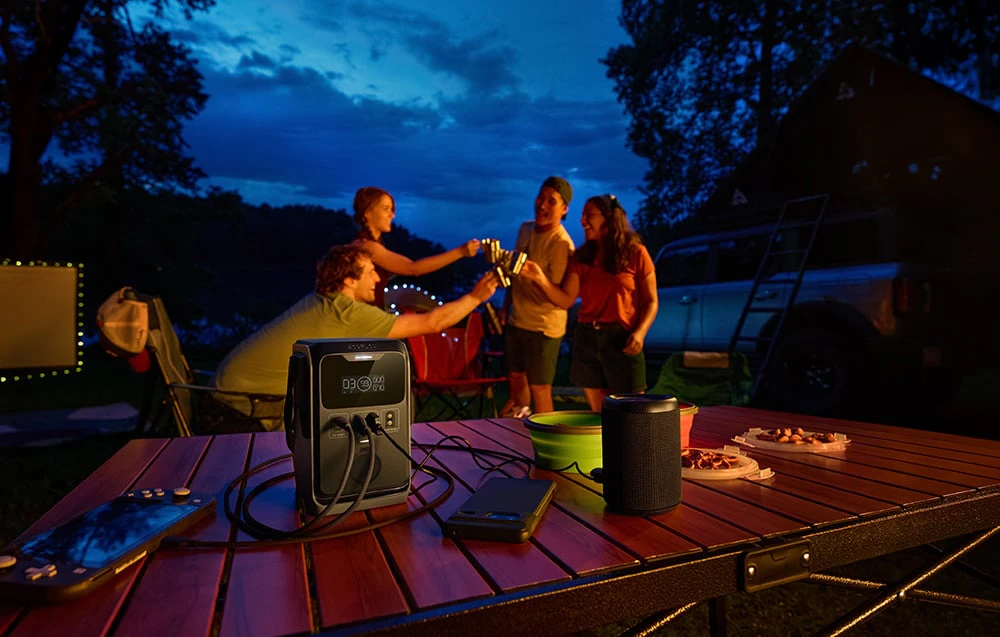 Un grup de oameni toastează seara la o masă de camping cu o stație electrică și un difuzor