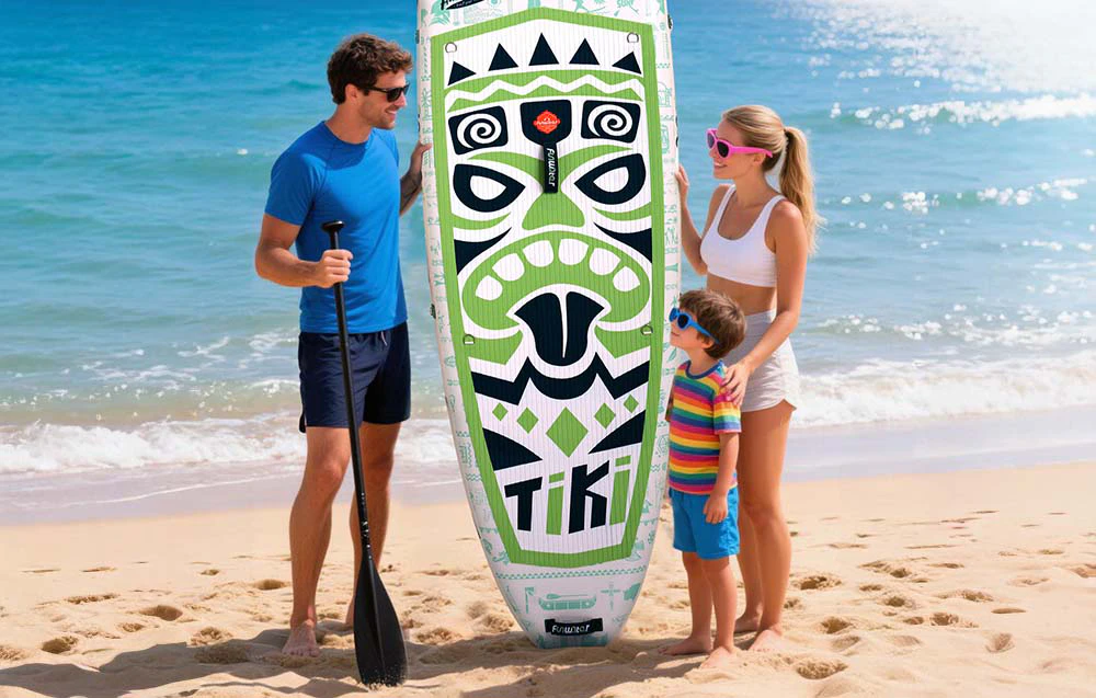 O familie zâmbitoare cu un copil stă pe plajă ținând o placă de surf și o vâslă, cu apă în spatele lor
