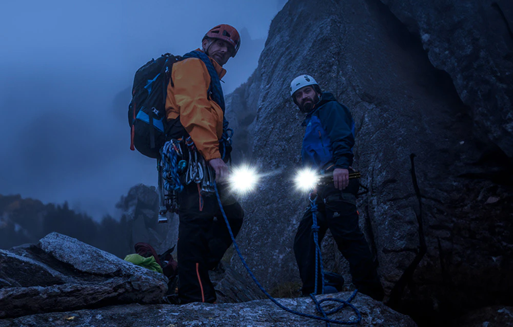 Doi alpiniști cu lanterne în mâini stând lângă stânci la amurg