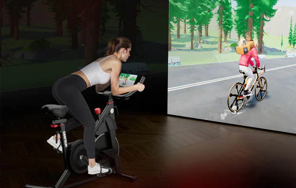 Femeie care se antrenează pe o bicicletă staționară în timpul unei curse virtuale afișate pe un ecran mare