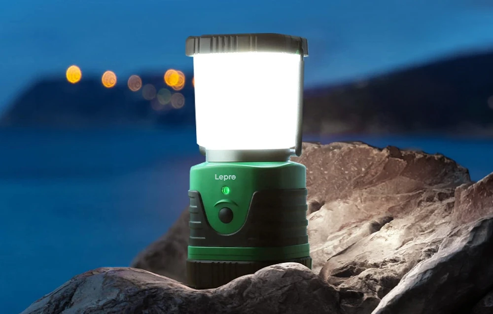 O lampă de camping luminează stâncile cu o lumină puternică; în fundal se văd lumina mării și a portului.
