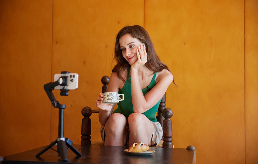 O femeie stă la masă, înregistrând un videoclip cu un smartphone montat pe un gimbal în timp ce bea cafea.